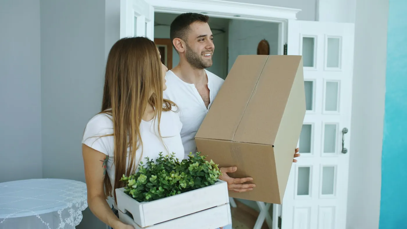 Twee mensen dragen verhuisdozen en een plantenbak hun nieuwe woning binnen